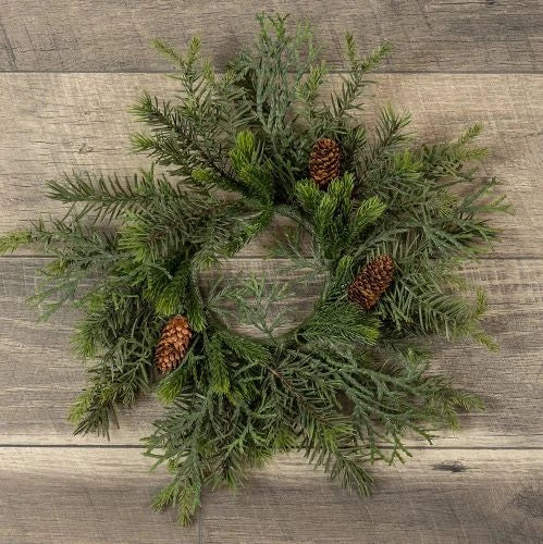 A rustic pine wreath made of green evergreen branches with three brown pinecones, displayed on a weathered gray-brown wooden plank surface.