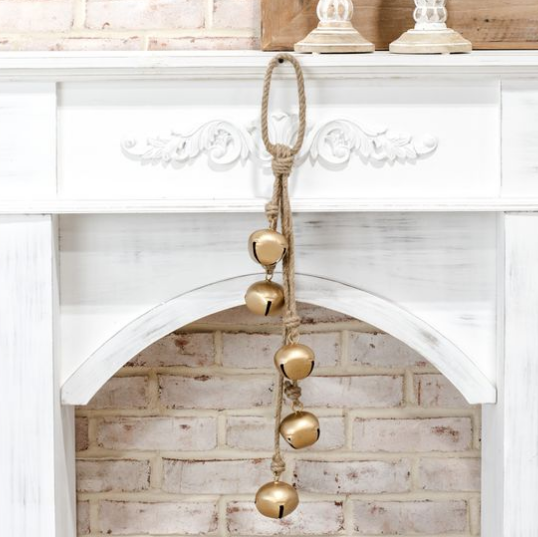 A rustic rope hanger with five gold-toned bells hanging in front of a white fireplace mantel with a brick interior.