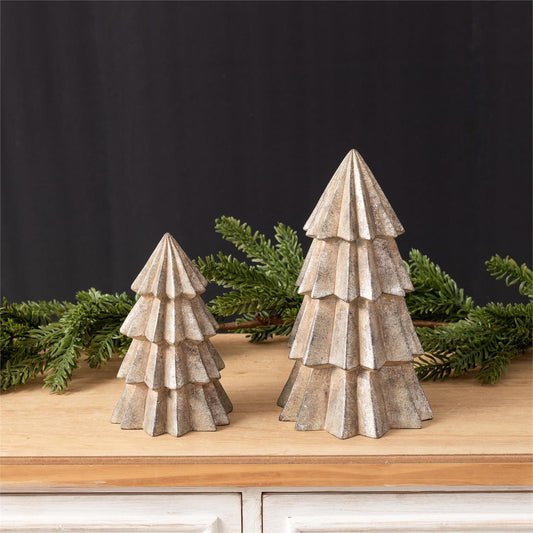 Two rustic, tiered metal Christmas tree decor in a distressed silver finish, displayed on a wooden surface with green pine branches against a dark background.