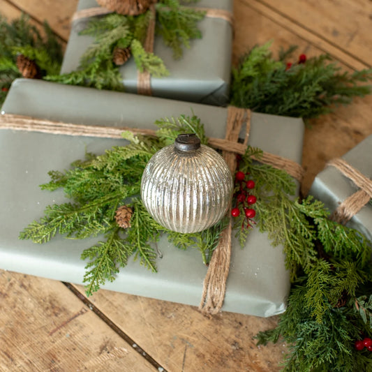 A silver ribbed glass Christmas ornament with a dark cap rests on a sage green wrapped gift box decorated with green pine branches, burlap ribbon, and small red berries, set on a rustic wooden table.