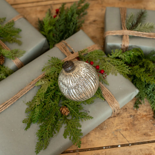 Three wrapped Christmas gifts in sage green paper with natural jute twine, decorated with green pine branches, small red berries, and a silver ribbed ornament, arranged on a rustic wooden surface.