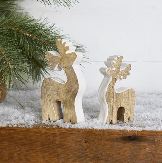 Two small natural wood reindeer figurines standing on a white faux-snow surface with pine branches at the top left.