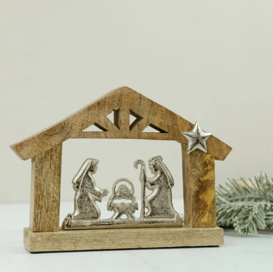 A small wooden nativity scene display with a silver-toned metal figure set inside a rustic house-shaped frame, shown on a light neutral background with a hint of greenery at the side.