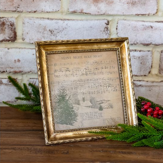 A square vintage-style framed print with a distressed gold frame, featuring sheet music reading 'Silent Night Holy Night' and a small tree illustration, displayed on a wooden surface against a white brick wall with greenery and red berries.