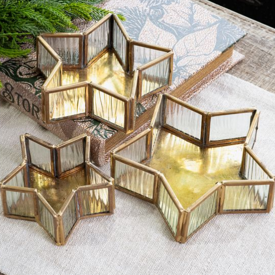 Three star-shaped glass and metal candle holders with gold-toned frames, arranged on a textured fabric surface with decorative books and greenery in the background.