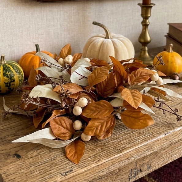 An autumn-themed table centerpiece with faux pumpkins, dried leaves, acorns, and candles on a rustic wooden table, with a large ornate mirror and books in the background.