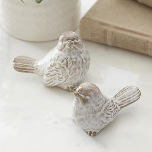 Two white with brown shading ceramic bird figurines farmhouse art on a light surface with a book and vase in the background.