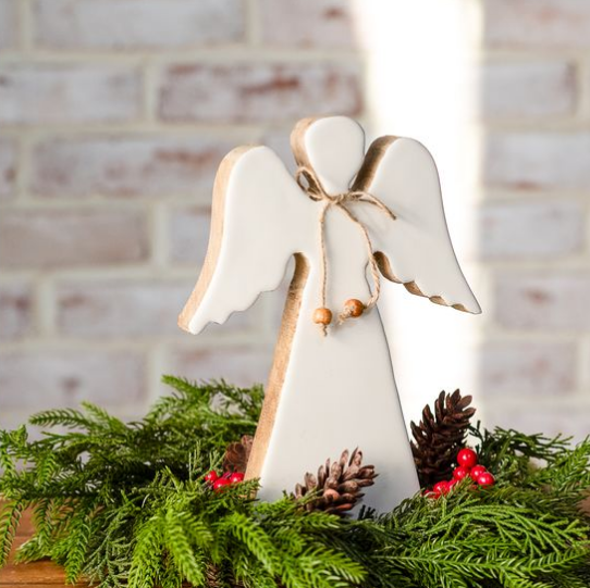 A white wooden angel figurine with natural wood edges, a jute bow, and small wooden beads, displayed in a holiday wreath with pinecones and red berries against a light brick wall background.