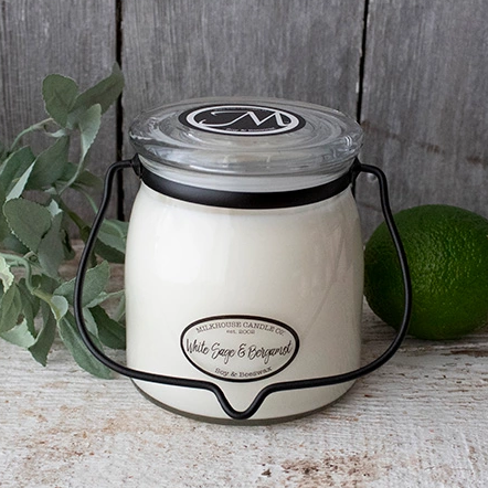 A white jar candle with a black lid and black metal handle, labeled 'White Sage & Bergamot', set against a rustic background with green foliage and a lemon.